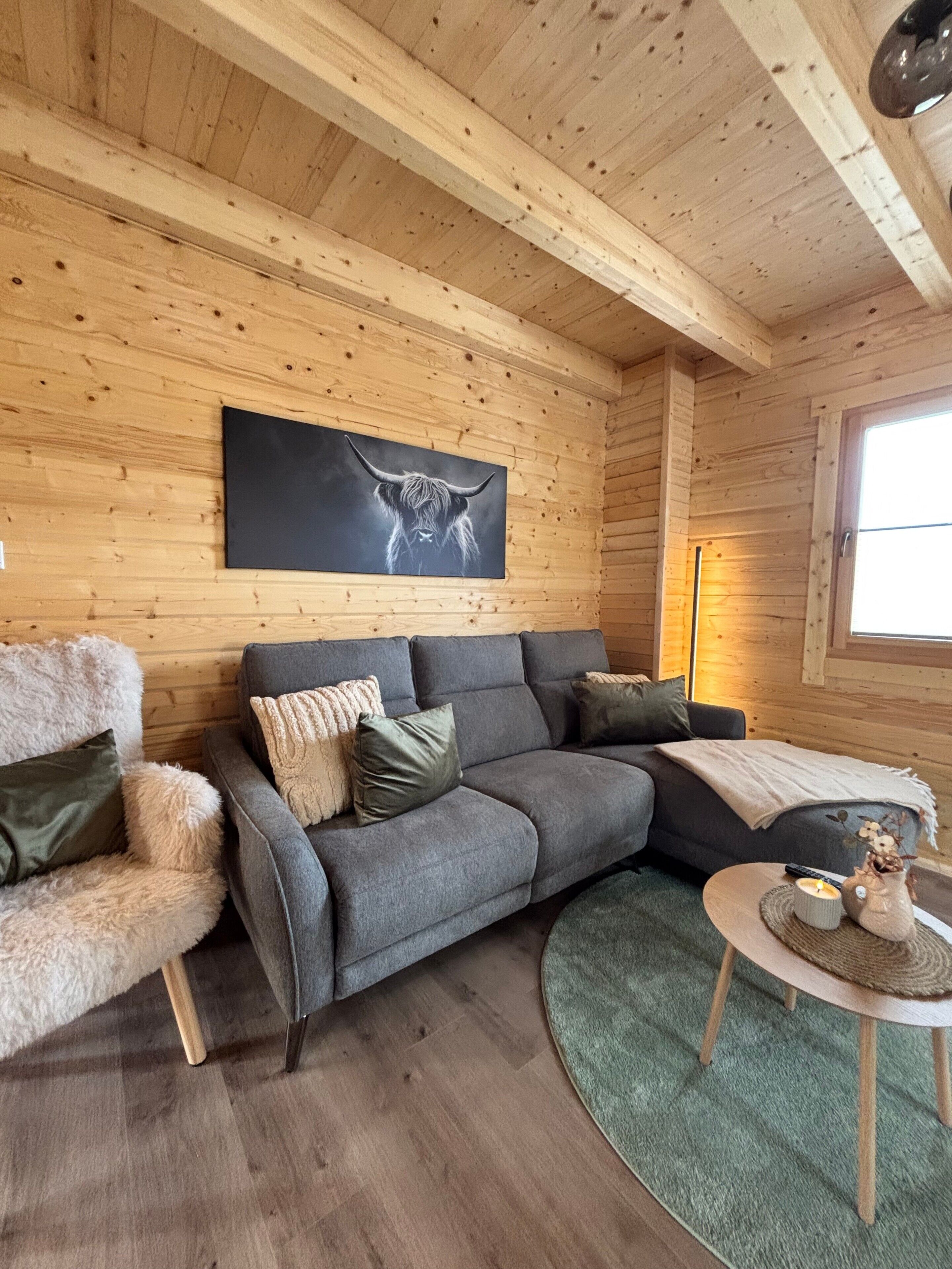 A cozy cabin interior with a gray sofa, a furry chair, a wooden ceiling, and a large window.