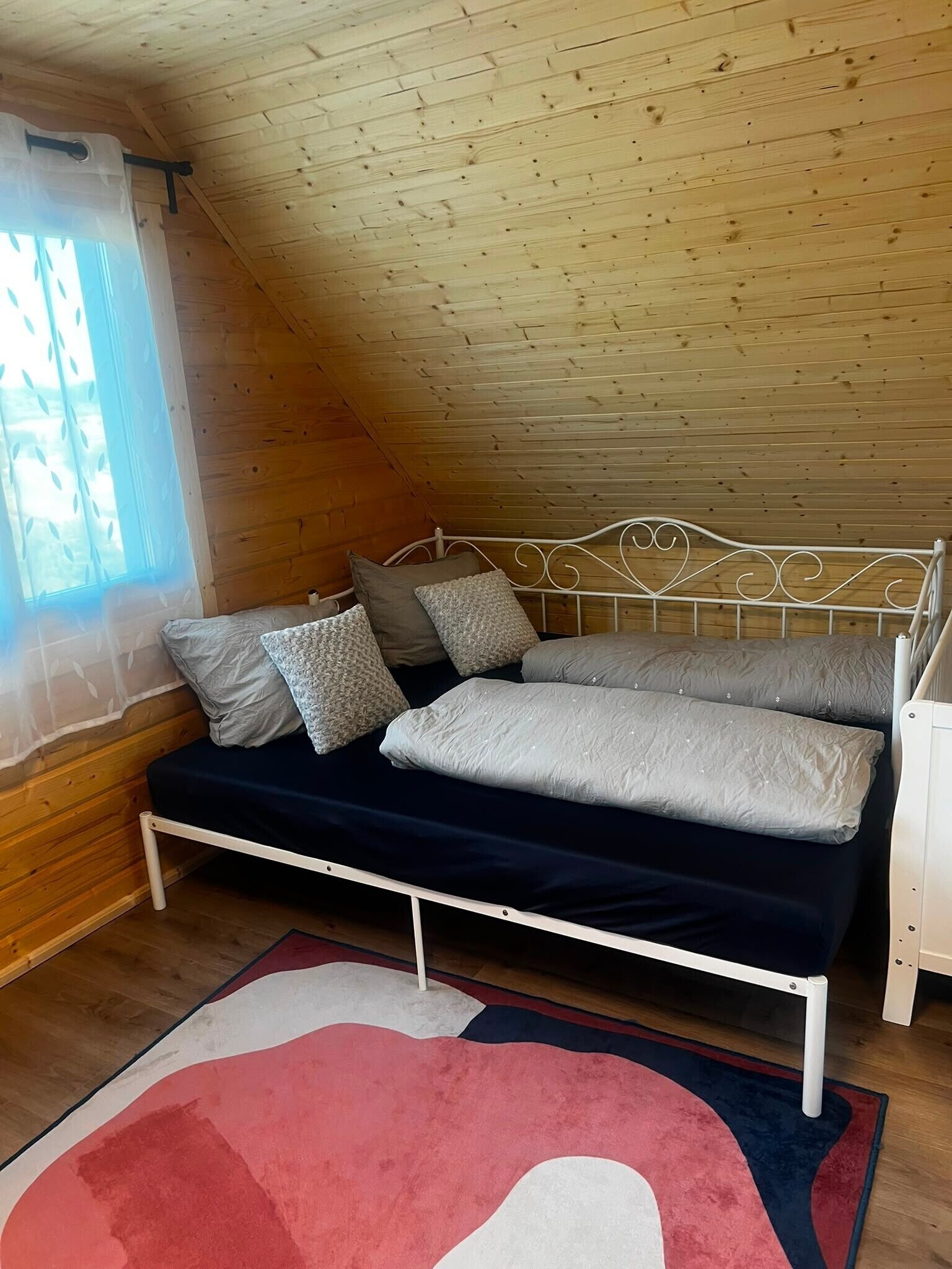 Une chambre à coucher aménagée dans un espace mansardé, avec un plafond en pente, un cadre de lit en métal et une grande fenêtre ornée de rideaux légers.