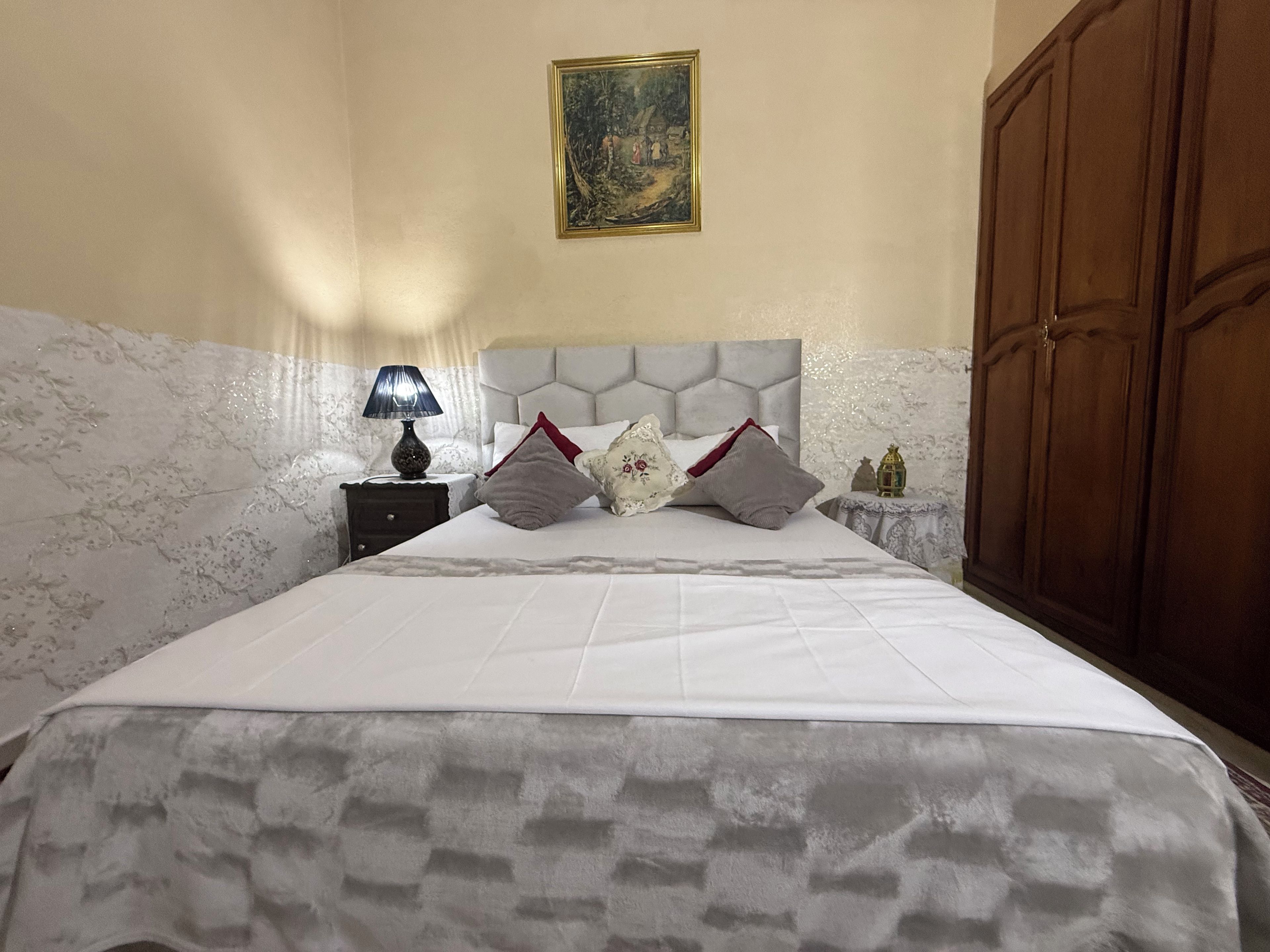 A neatly made bed with a headboard, a bedside table with a lamp, and a painting on the wall.