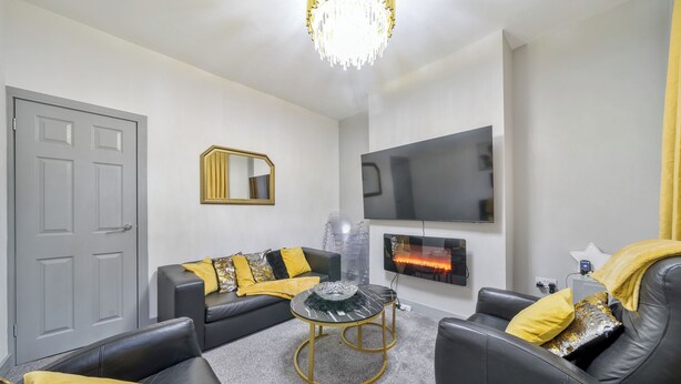 A living room with a grey door, black leather sofa, a round coffee table, a flat-screen TV mounted on the wall, and a chandelier.