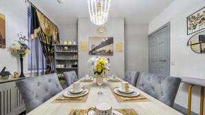 A dining room with a long table set for six, grey upholstered chairs, a chandelier, a clock, and a cityscape painting on the wall.