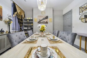 Une salle à manger avec une longue table dressée pour six personnes, des chaises à revêtement gris, un lustre, une horloge et un tableau représentant un paysage urbain accroché au mur.