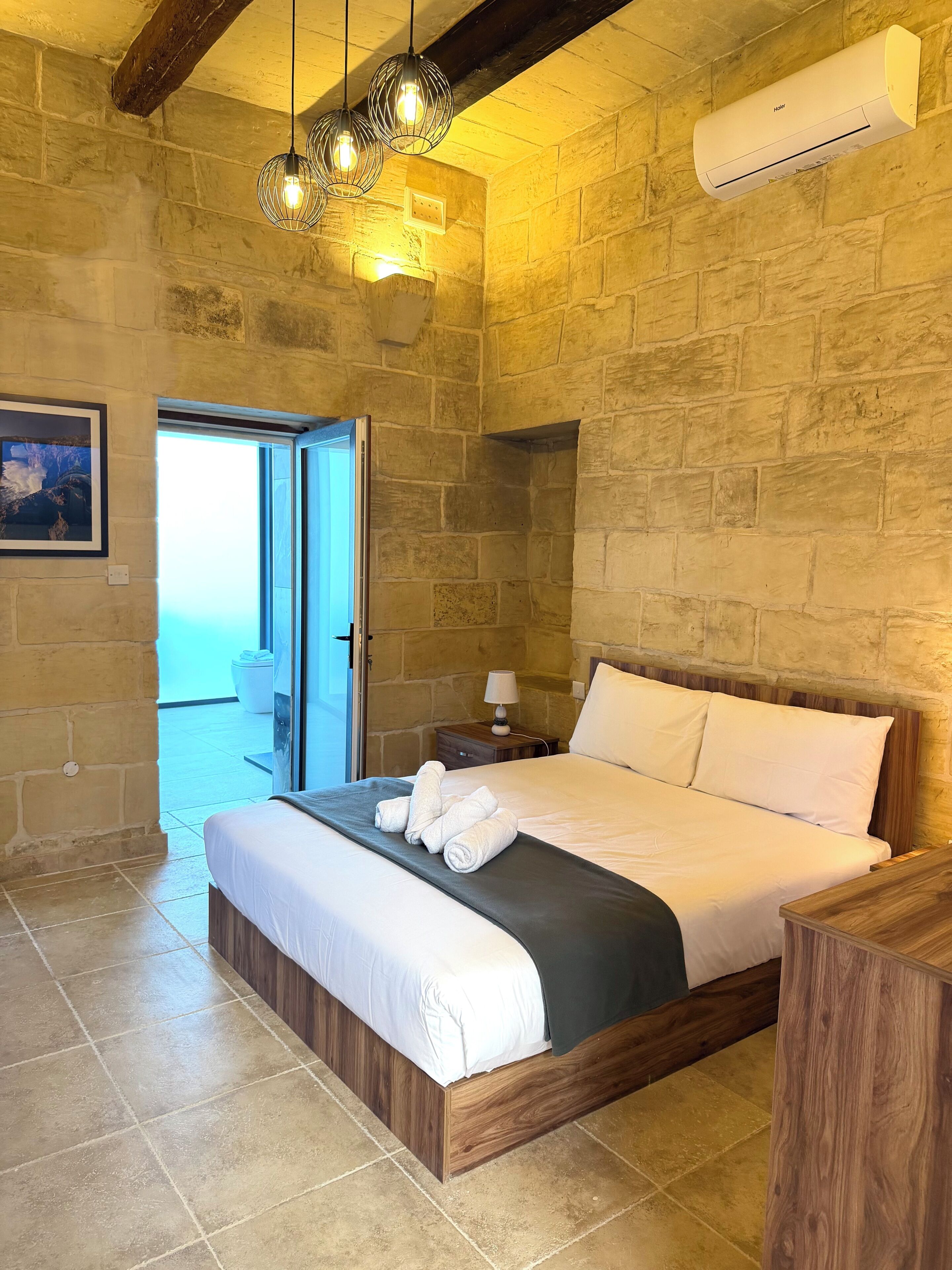 A bedroom with a stone wall, a bed with white linens, a wooden headboard, a nightstand with a lamp, and a view through an open door.