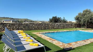 A swimming pool with a stone border, surrounded by artificial grass, and a stone wall in the background.