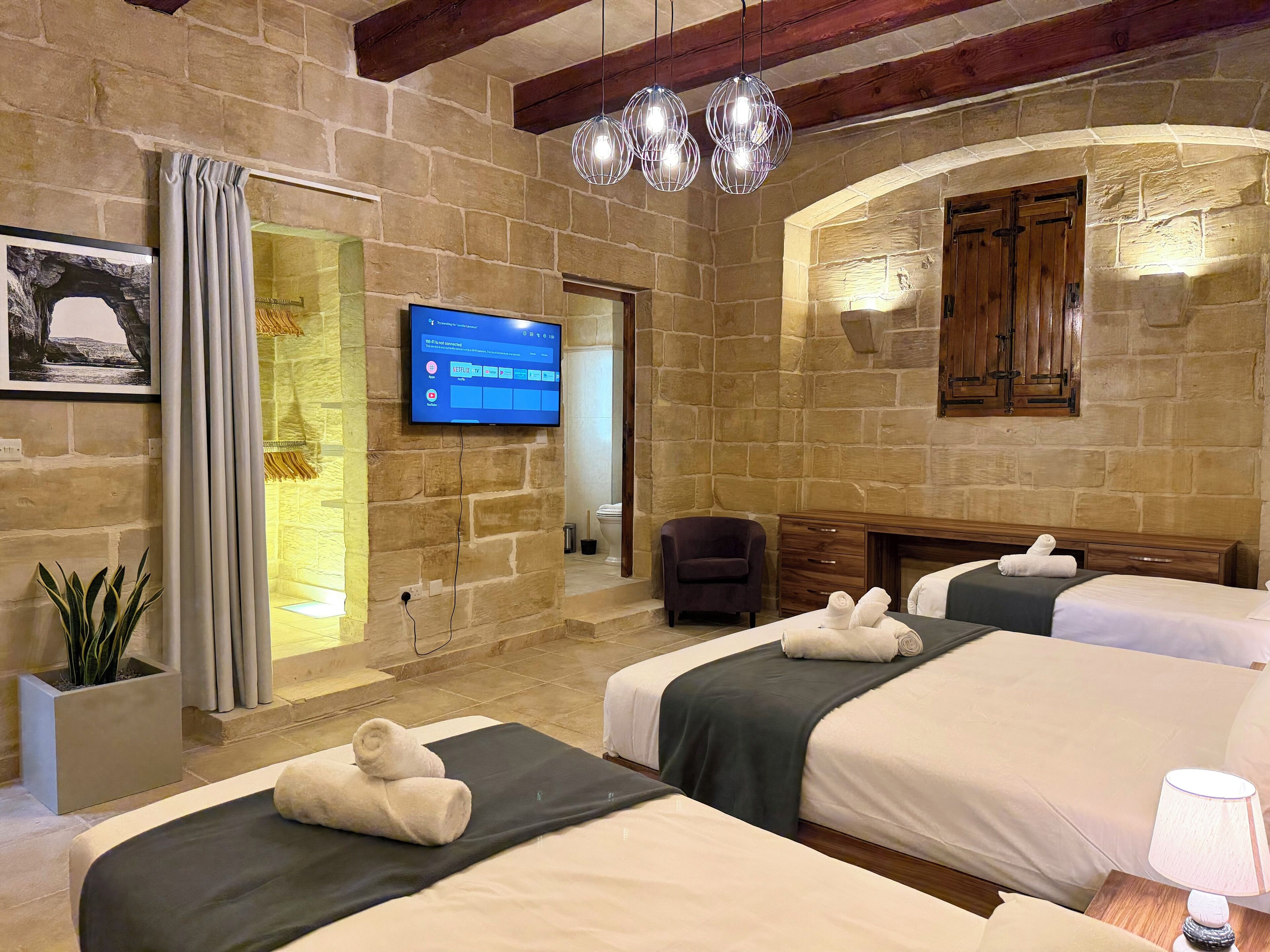 A hotel room with stone walls, a flat-screen TV, two beds with towels, a wooden cabinet, and a potted plant.
