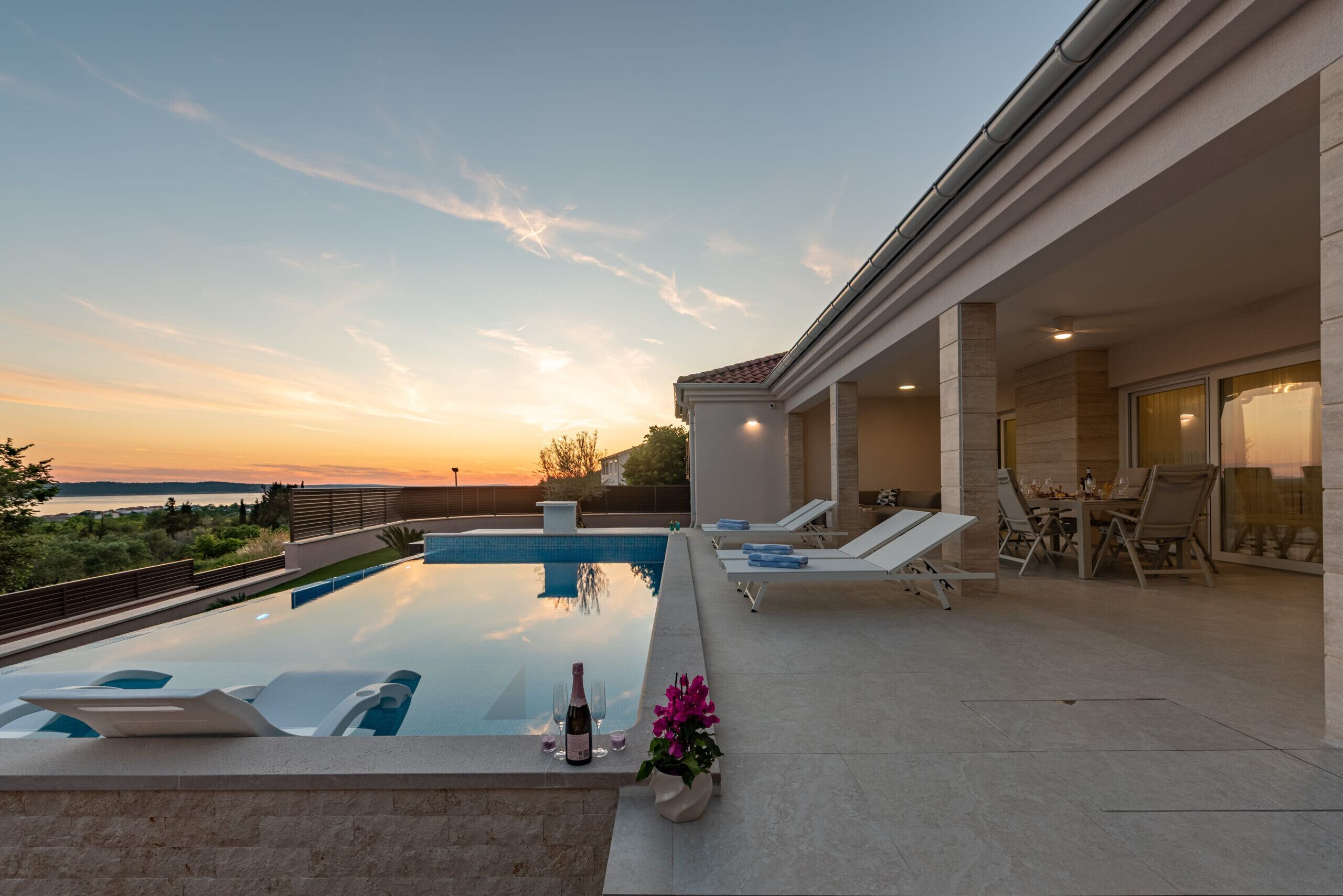 A pool with lounge chairs, a table with chairs, and a view of the sunset.