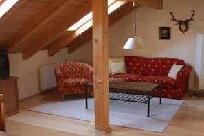 Ein Loft-Zimmer mit Holzbalkendecke, einem rot gemusterten Sofa, einem gemusterten Sessel, einem Glastisch und einem aufgehängten Damhirschschädel an der Wand.