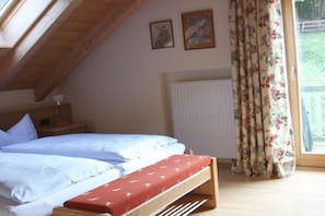 Ein Schlafzimmer mit schrägemaßtem Dach, einem Bett mit weißer Bettwäsche, einer hölzernen Sitzbank, einem Heizkörper und floralen Vorhängen.