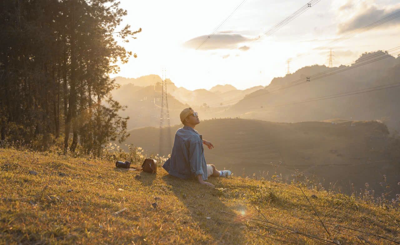 A person sitting on a grassy hill with a backpack and a bag, overlooking a valley with power lines and trees.