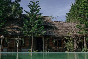 Ein Holzhaus mit einem Strohdach, umgeben von Bäumen und einem Schwimmbecken.