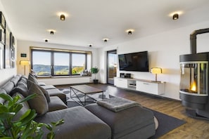 A modern living room with a sofa, TV, fireplace, and large windows. - Holiday home in Grafenau with swimming pool (Grafenau)