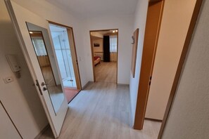 Ein Flur mit Holzboden, einer Tür mit Glasfenster und einem Raum mit Bett und Vorhängen im Hintergrund.