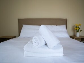Ein ordentlich bezogenes Bett mit weißen Leinen und einem aufgerollten Handtuch darauf, ein hölzerner Kopfteil und ein Nachttisch mit einer Vase gelber Blumen.