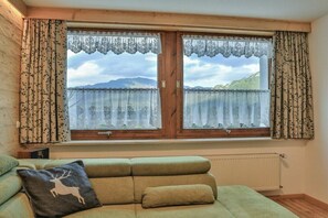 Ein Zimmer mit einem grünen Sofa, einem Fenster mit Spitzenvorhängen und Blick auf Berge.