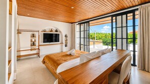 A bedroom with a wooden bed, a TV, a balcony with a view of greenery, and a wardrobe with shelves. - Enclosed Modern 3BR, with Private Pool & Sunken Lounge (Badung)