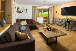 Ein Wohnzimmer mit braunem Sofa, einem Glastisch mit Früchten, einer Steinwand und einem an der Wand montierten Fernseher.