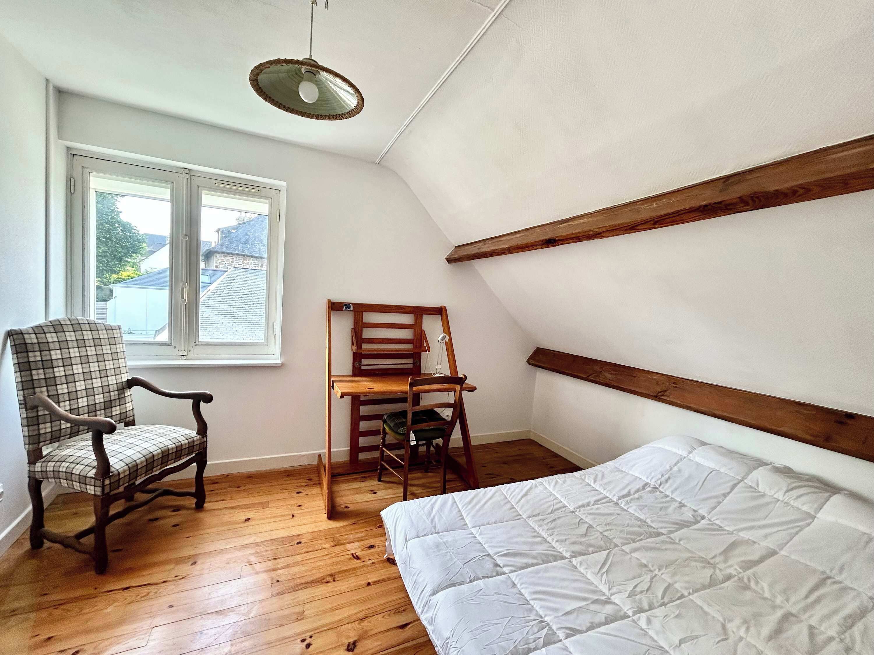 Ein Zimmer mit Holzboden, einem Bett mit weißer Decke, einem Karo-Sessel, einem Holzstuhl und einem Fenster mit Blick auf Gebäude.