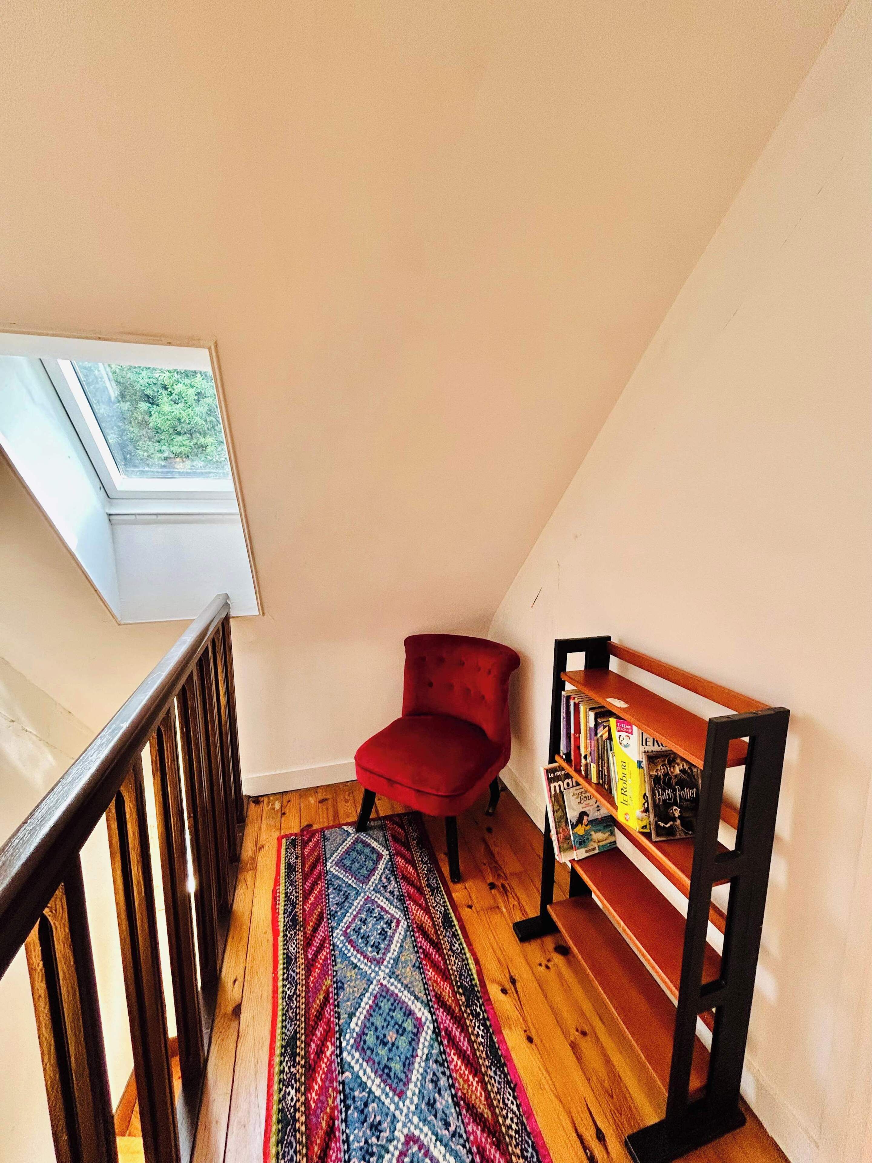 Una habitación con piso de madera, una silla roja, una estantería y un ventanal con vistas a la vegetación.