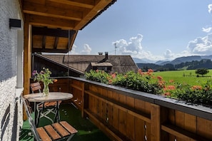 A wooden balcony with a table and chairs, a flower vase, and a view of green fields and mountains. - Holiday apartment with 3 bedrooms (Oberstdorf-Schöllang)