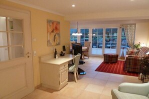 Ein Wohnzimmer mit einer Couch, einer Essecke mit Tisch und Stühlen, einem Schreibtisch mit Computer und einem großen Fenster mit Blick ins Freie.