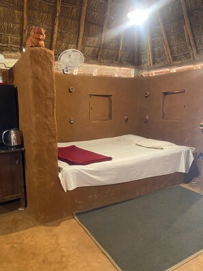 Una habitación rústica con una cama, un ventilador, una escultura de cerdo y un techo de paja.