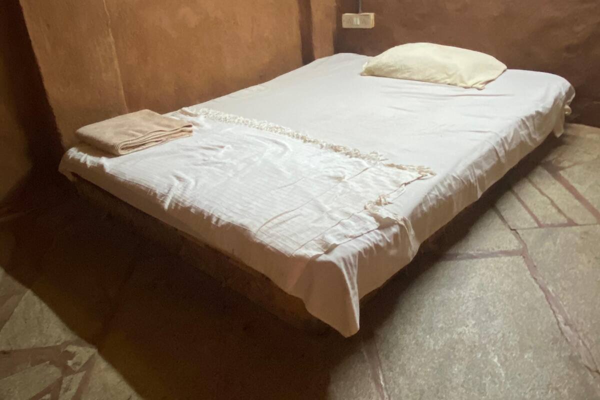 Ein Einzelbett mit weißer Bettdecke und beiger Decke, ein kleines Fenster mit Metallrahmen sowie eine an der Wand befestigte Leuchte.