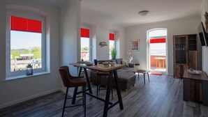 Un vaste espace de vie comprenant une table à manger, des chaises, un canapé et un téléviseur. De grandes fenêtres équipées de stores rouges offrent une vue sur l’extérieur.