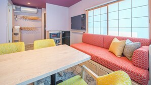 Una habitación con una mesa de madera, sillas con estampado amarillo, un sofá rojo con estampado y una televisión, además de una ventana con persianas.