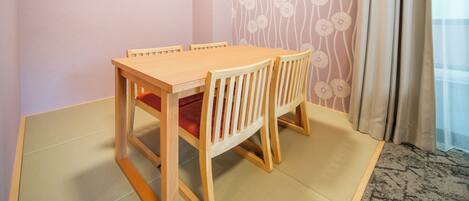 Una habitación con una mesa de comedor de madera, sillas, esteras de tatami, un papel tapiz estampado y una ventana con cortinas.