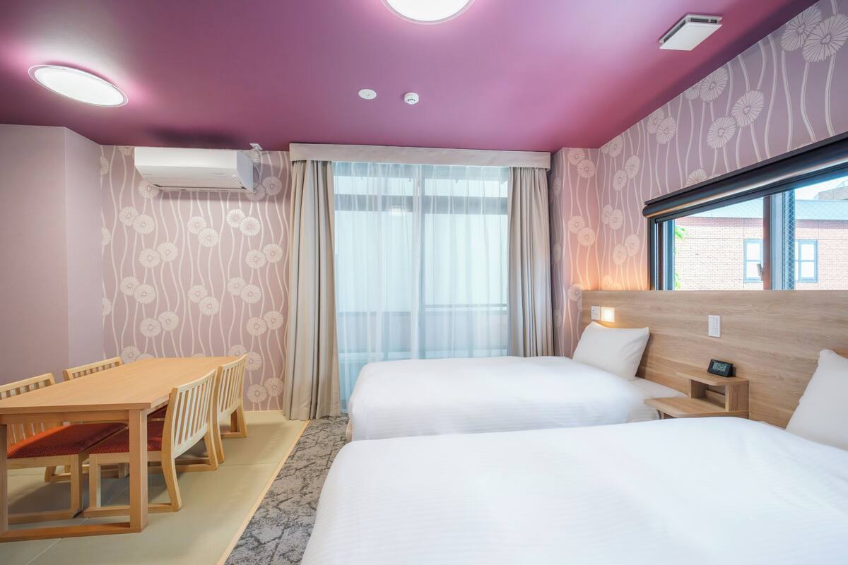 A hotel room with a wooden table, two beds, a window with curtains, and a ceiling with recessed lighting.