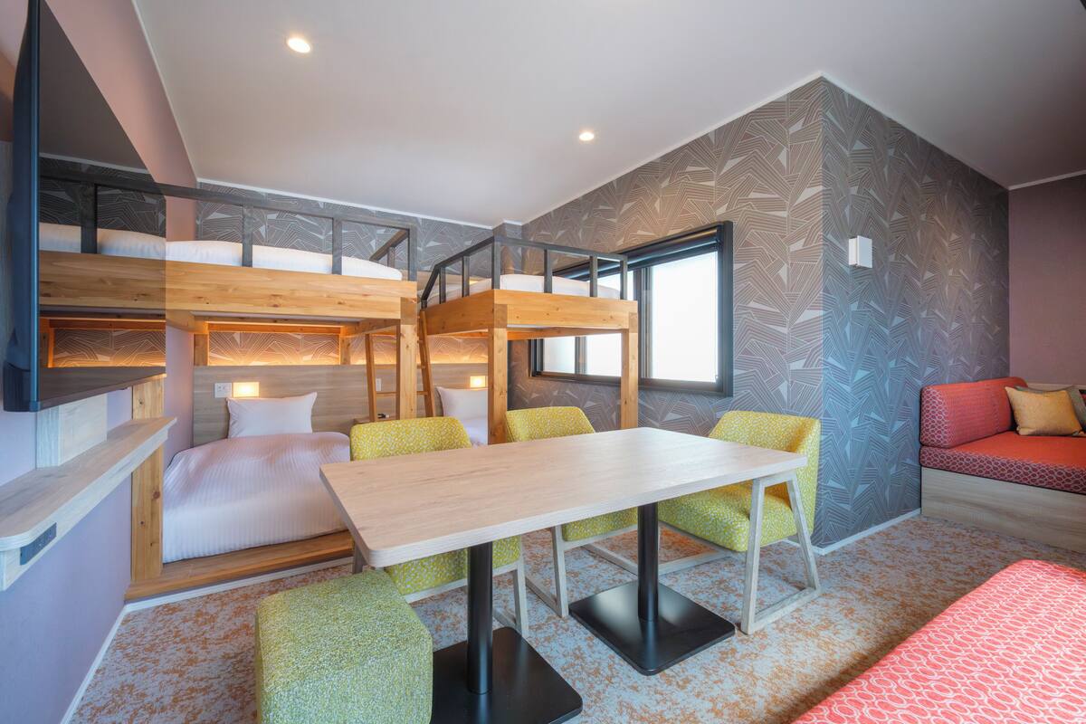 A room with bunk beds, a table, and chairs, with a patterned wall and a window.