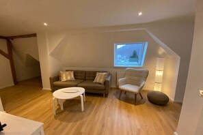 Ein Loft-Zimmer mit Holzboden, einer Couch, einem Sessel, einem kleinen Tisch und einem Fenster mit Blick auf eine verschneite Landschaft.