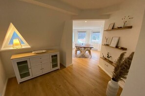 Ein Zimmer mit Holzboden, einem weißen Schrank mit Glastüren, einem Tisch mit Stühlen und einem Fenster mit einer dreieckigen Leuchte.