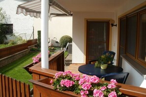 Eine Terrasse mit einer Holzbank, einem Tisch mit einem Blumentopf und Blick auf einen Garten mit Hecke und Vogelhaus.