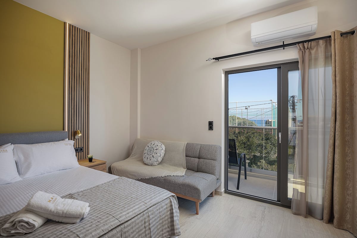 A modern hotel room with a bed, a sofa, a balcony with a view, and an air conditioning unit.