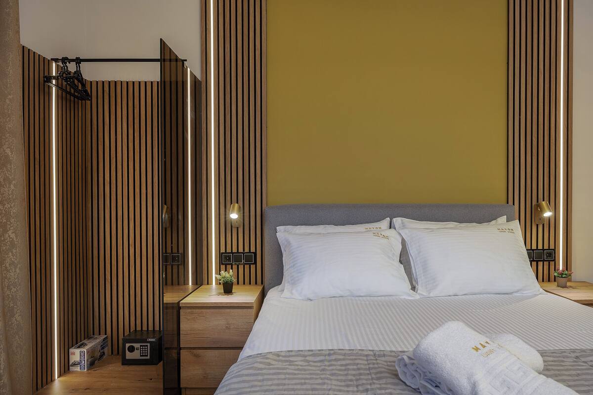 A hotel room with a bed, bedside tables, a nightstand, a wall-mounted lamp, and a wooden panel design.