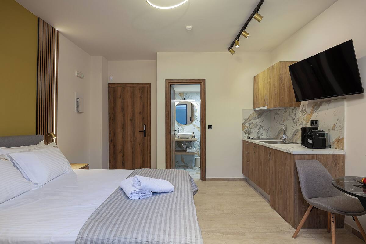 A hotel room with a bed, a small desk, a chair, a TV mounted on the wall, and a kitchen area with a sink and countertop.