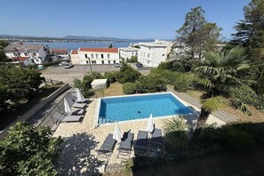 Ein Swimmingpool mit Liegestühlen und Sonnenschirmen, umgeben von Grünflächen und Gebäuden.