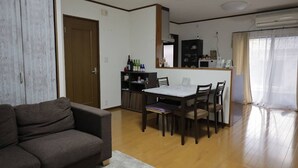 Un salon avec un canapé, une table à manger avec des chaises, un coin cuisine avec un réfrigérateur et des étagères, et une grande fenêtre avec des rideaux.