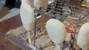 Un support métallique sur lequel sont disposés plusieurs objets blancs, peut-être des aliments, et qui est placé au-dessus d’un feu.