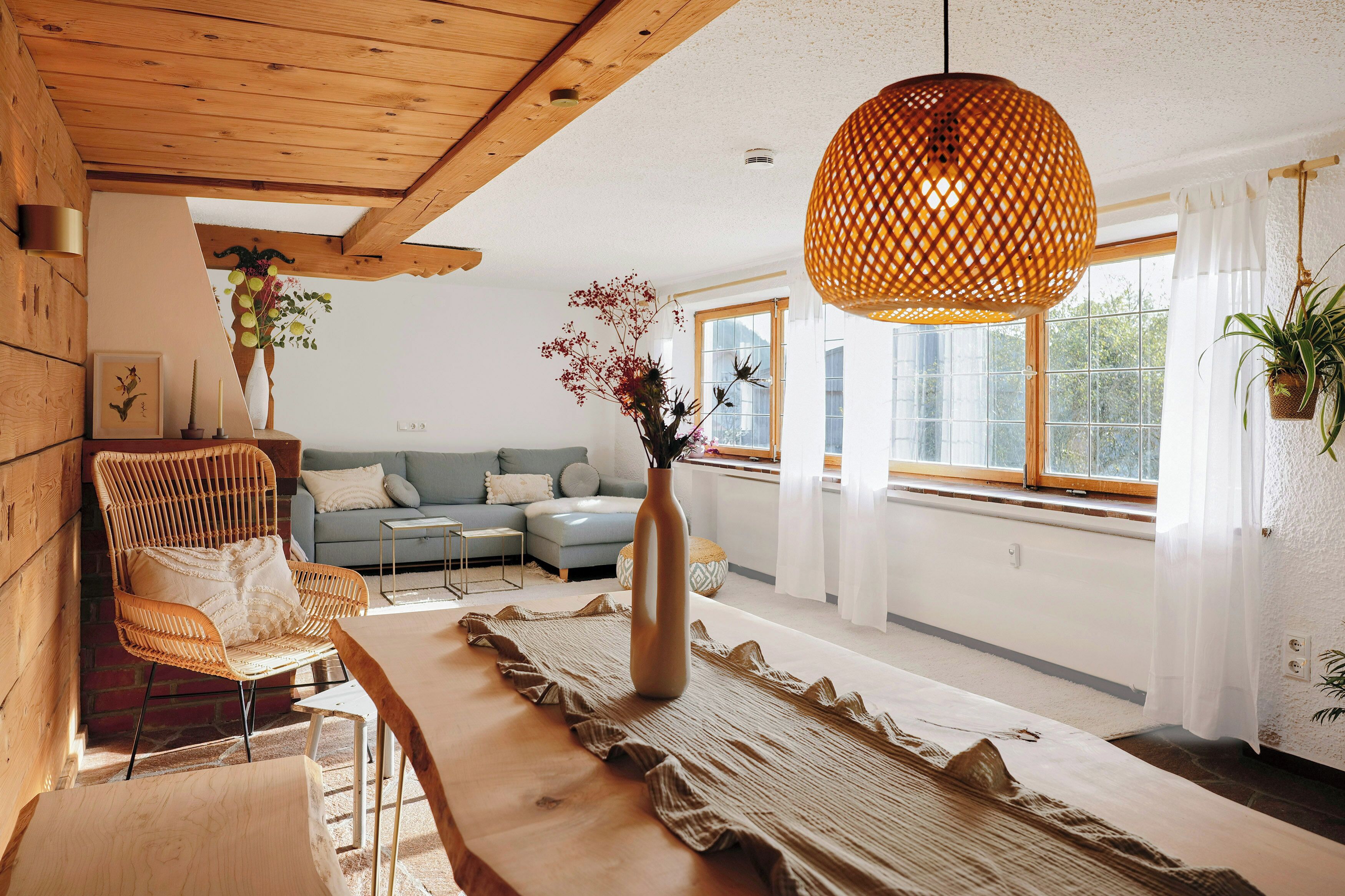 Uma sala de estar moderna com teto de madeira, luminária pendente trançada, conjunto de sofá, mesa de centro e um vaso com flores.