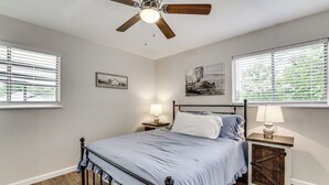Un dormitorio con cama, dos mesitas de noche, ventilador de techo y dos ventanas con persianas.