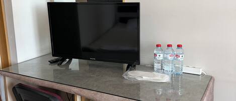 Habitación de hotel con escritorio de vidrio, monitor negro, mini refrigerador y una silla.