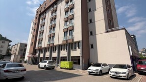 Un edificio de varios pisos con fachada plana, varios balcones y un primer piso empedrado con coches aparcados.