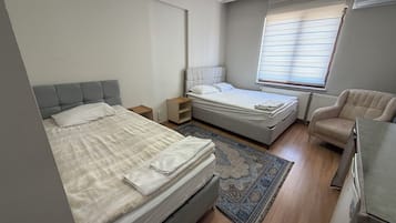 A bedroom with two beds, a small bedside table, a chair, a radiator, and a window with blinds.