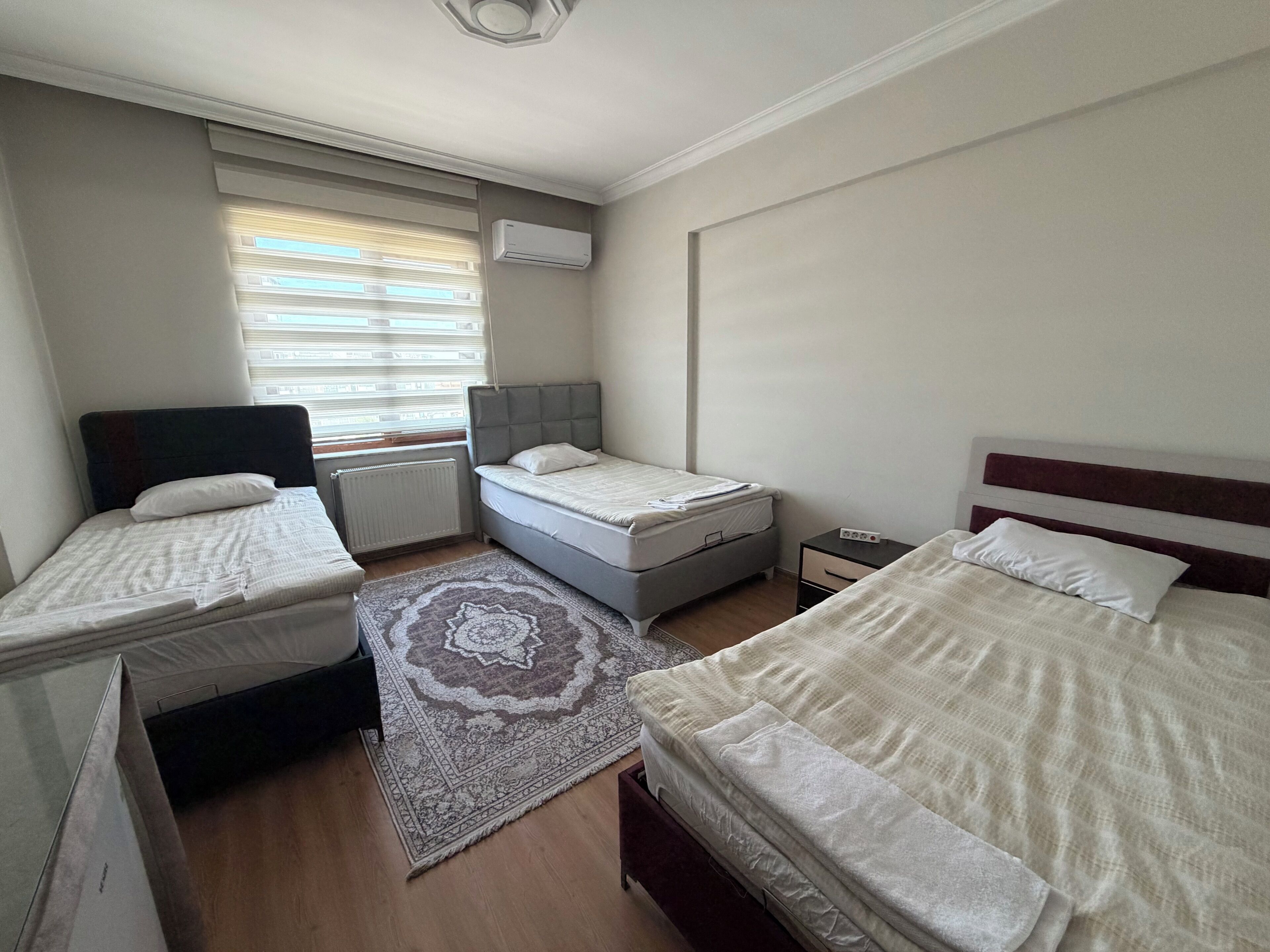 A hotel room with two beds, a window with blinds, an air conditioning unit, a bedside table with a lamp, and a rug.