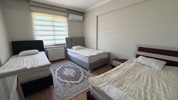 A hotel room with two beds, a window with blinds, an air conditioning unit, a bedside table with a lamp, and a rug.