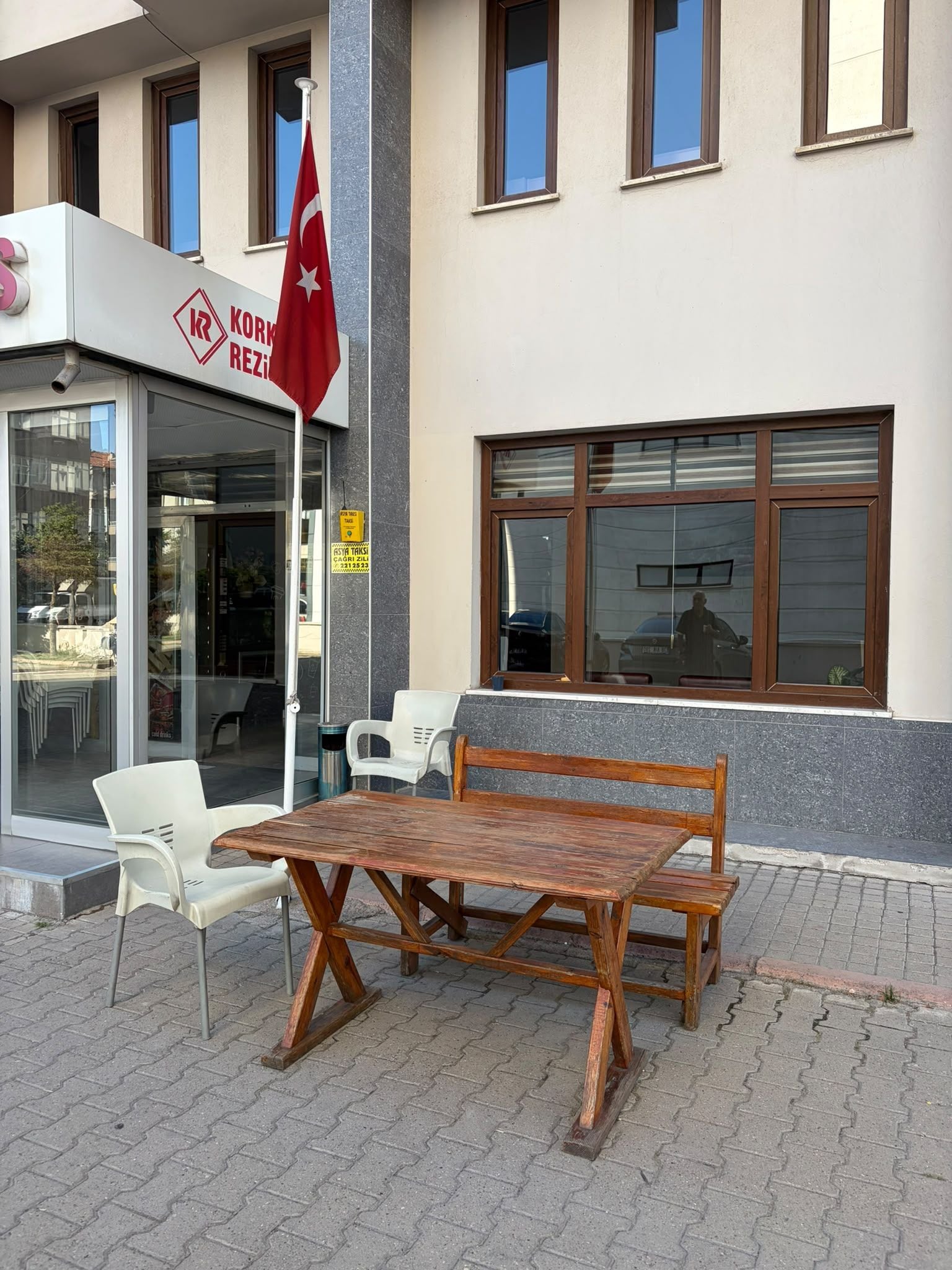 A building with a sign reading 'KORREZI' and a Turkish flag. There is a wooden bench and two white plastic chairs outside.