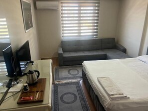 Ein Hotelzimmer mit einem Bett, einem Sofa, einem Schreibtisch mit Computer, einer Kaffeemaschine und einem Fenster mit Jalousien.