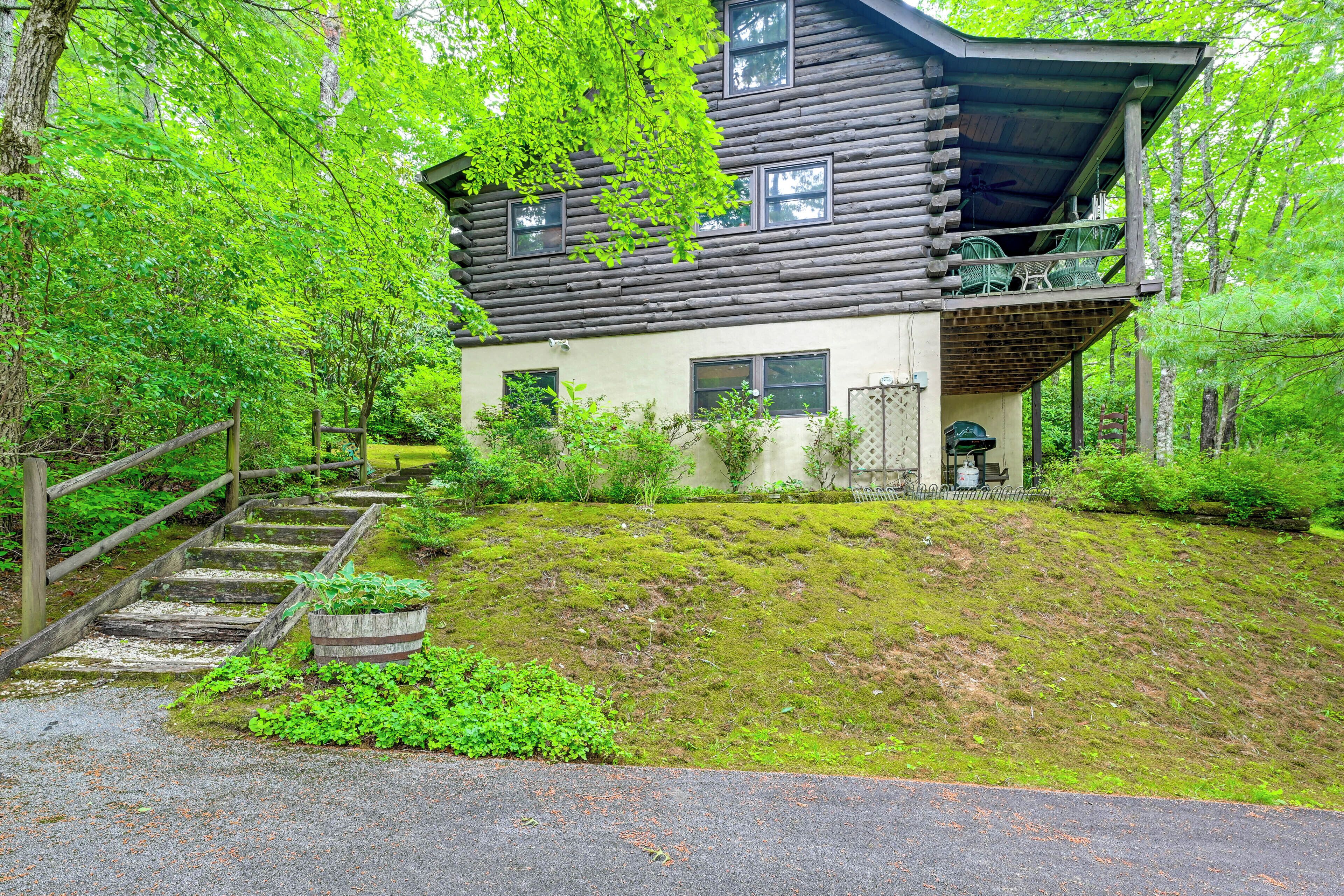 A log cabin with a balcony, surrounded by lush greenery and a wooden staircase leading up to the entrance.